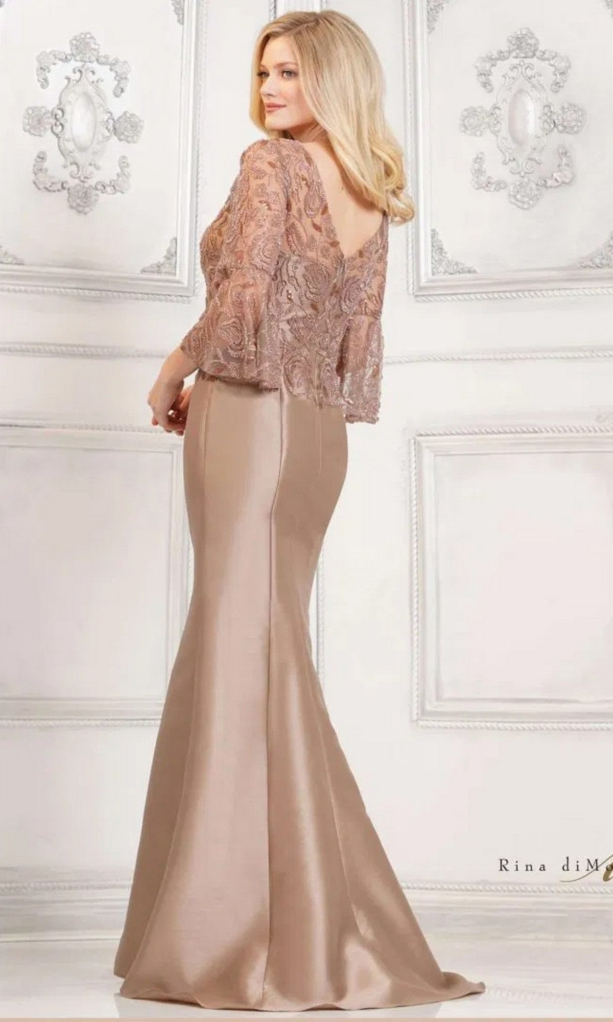 A woman wearing the Rina Di Montella RD2939 long formal dress with a beaded bodice and flared satin skirt stands in a decorated white room.